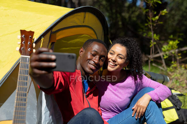 Feliz casal diversificado sentado na tenda e tomando selfie no campo. saudável, estilo de vida ao ar livre ativo e tempo de lazer. — Fotografia de Stock
