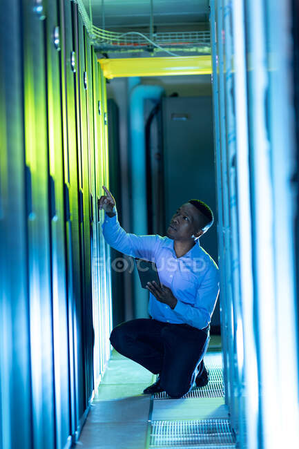 Técnico de computadora afroamericano masculino usando tableta trabajando en la sala de servidores. tecnología de redes digitales de almacenamiento y comunicación de información. - foto de stock