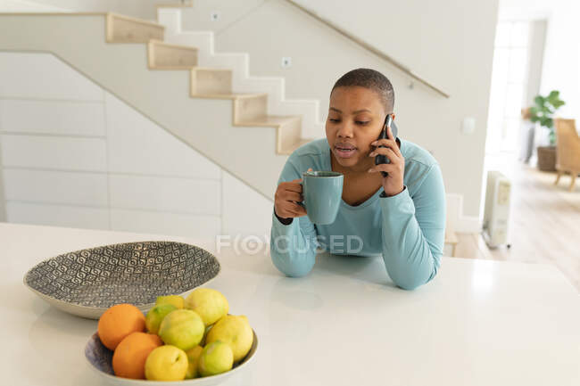 Afro-americano plus size mulher fazendo chamada e beber café na cozinha. estilo de vida, lazer, passar tempo em casa com a tecnologia. — Fotografia de Stock