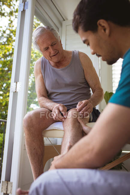 Fisioterapeuta masculino biracial tratando perna de paciente masculino sênior na clínica. cuidados de saúde seniores e tratamento de fisioterapia médica. — Fotografia de Stock