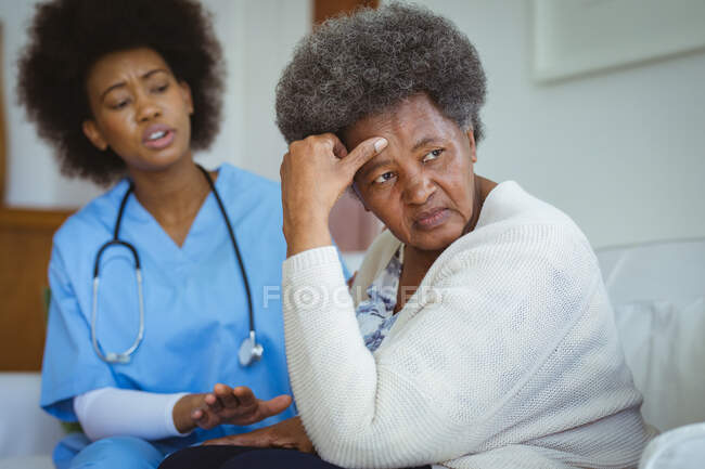 African american female doctor talking with sad senior female patient at home. healthcare and lifestyle during covid 19 pandemic. — Stock Photo
