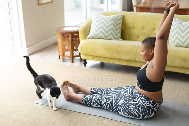 Felice afroamericano plus size donna che pratica yoga su tappetino a casa con il gatto. fitness e stile di vita sano e attivo. — Foto stock