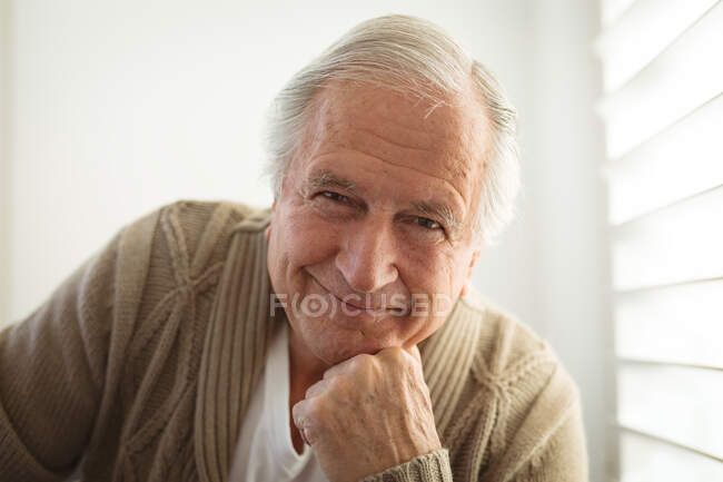 Portrait of smiling senior caucasian man looking at camera and touching his chin at home. spending time at home alone. — Stock Photo
