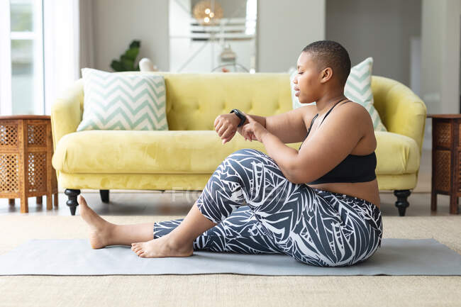Mujer afroamericana de talla grande practicando yoga en la esterilla, revisando smartwatch. fitness y estilo de vida saludable y activo. - foto de stock