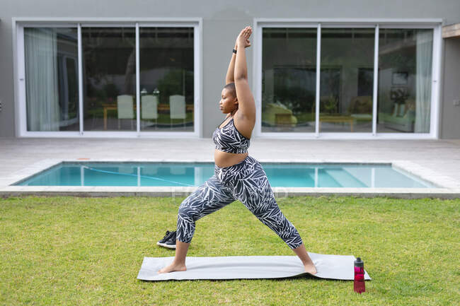 Focused african american plus size woman practicing yoga on mat in garden by swimming pool. fitness and healthy, active lifestyle. — Stock Photo
