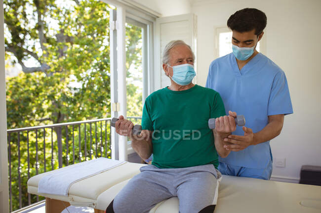 Physiothérapeute mâle naissante avec masque facial traitant les bras d'un patient masculin âgé à la clinique. soins de santé supérieurs et traitement de physiothérapie médicale. — Photo de stock