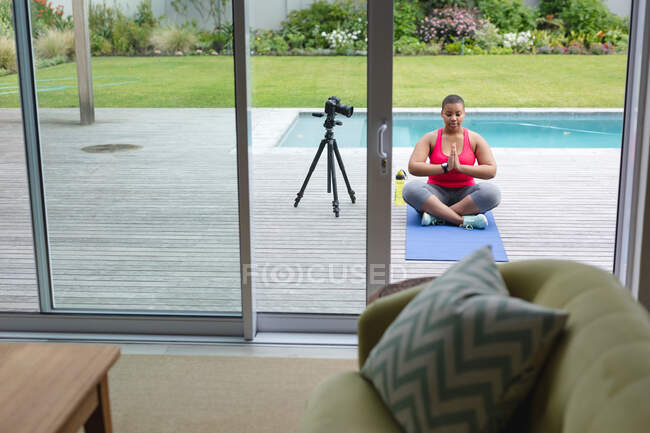 African american plus size woman in sports clothes sitting on mat and practicing yoga, making vlog. fitness and healthy, active lifestyle. — Stock Photo
