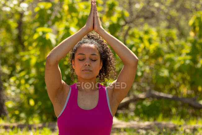 Femme biraciale relaxante pratiquant le yoga, assise et méditant à la campagne. mode de vie sain et actif en plein air et temps libre. — Photo de stock