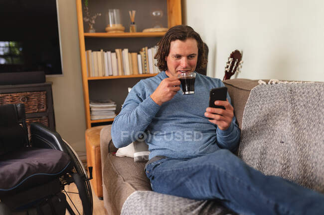 Caucasian disabled man drinking coffee and using smartphone sitting on the couch at home. disability and handicap concept — Stock Photo