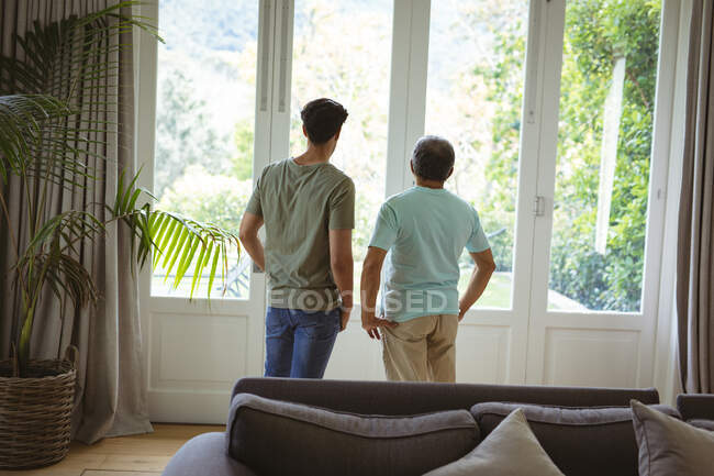 Hijo adulto birracial y padre mayor mirando por la ventana de la sala de estar. tiempo en familia en casa juntos. - foto de stock
