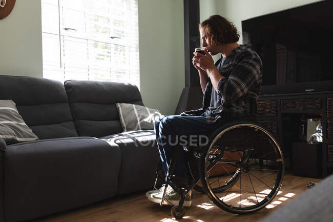Thoughtful caucasian disabled man sitting on wheelchair drinking coffee at home. disability and handicap concept — Stock Photo