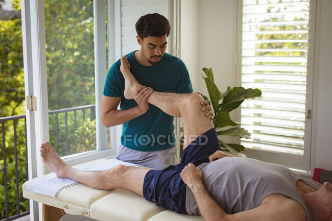 Біраціонний чоловічий фізіотерапевт, який лікує ногу старшого пацієнта чоловічої статі в клініці. лікувальна та медична фізіотерапія . — стокове фото