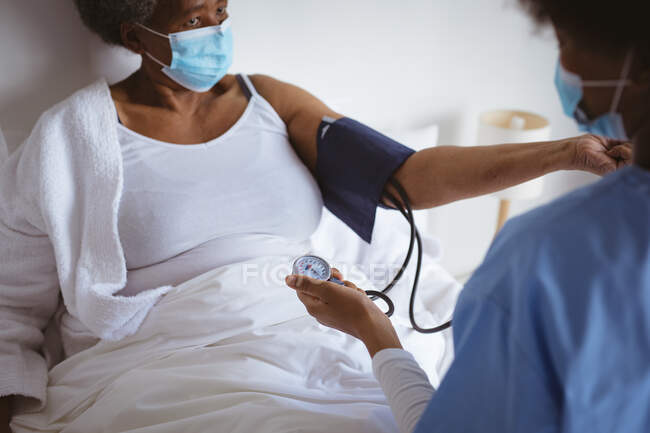 Médico afroamericano con mascarilla facial presionando a una paciente mayor en casa. salud y estilo de vida durante la pandemia de covid 19. - foto de stock