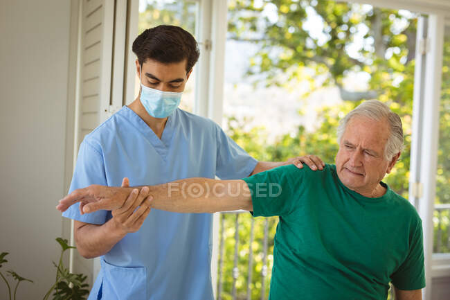 Бирасенсорный физиотерапевт-мужчина в маске для лица лечит руки пожилого пациента в клинике. медицинское и физиотерапевтическое лечение. — стоковое фото