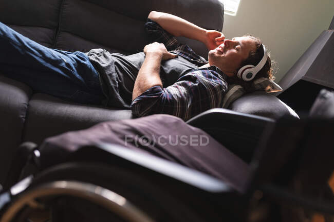 Hombre caucásico discapacitado que usa auriculares escuchando música mientras está acostado en el sofá de su casa. concepto de discapacidad y discapacidad - foto de stock