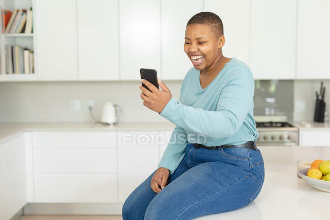 Feliz afro-americano plus size mulher ter chamada de vídeo no smartphone na cozinha. estilo de vida, lazer, passar tempo em casa com a tecnologia. — Fotografia de Stock