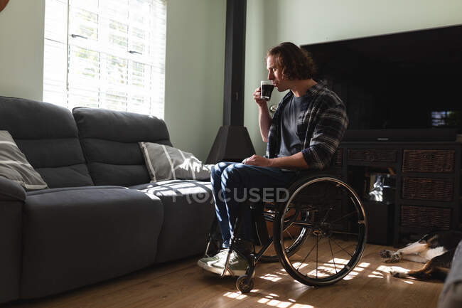 Homem caucasiano deficiente pensativo sentado em cadeira de rodas bebendo café em casa. conceito de deficiência e deficiência — Fotografia de Stock