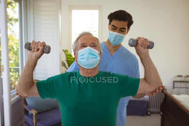 Biracial male physiotherapist with face mask treating arms of senior male patient at clinic. senior healthcare and medical physiotherapy treatment. — Stock Photo