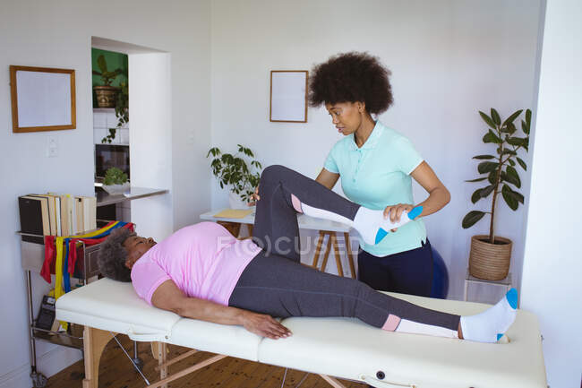 Африканская американка-физиотерапевт лечит ногу пожилой пациентки в клинике. медицинское и физиотерапевтическое лечение. — стоковое фото