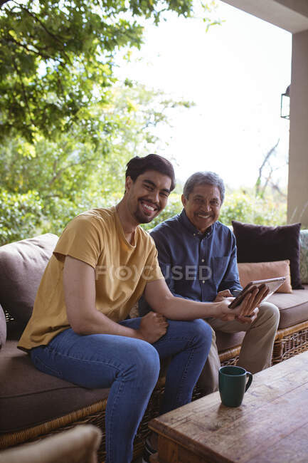 Retrato de hijo adulto birracial sonriente y padre mayor usando tableta. tiempo en familia en casa juntos. - foto de stock