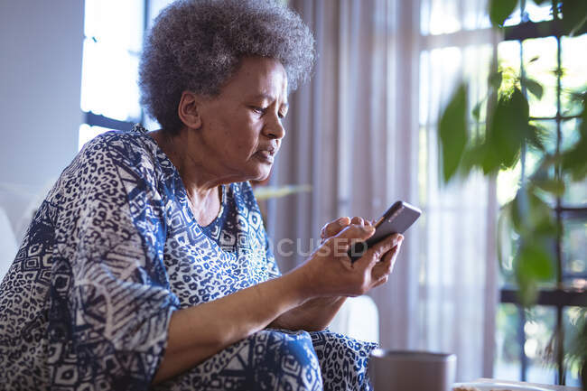 Donna anziana afroamericana seduta e utilizzando smartphone. passare del tempo a casa usando solo la tecnologia. — Foto stock
