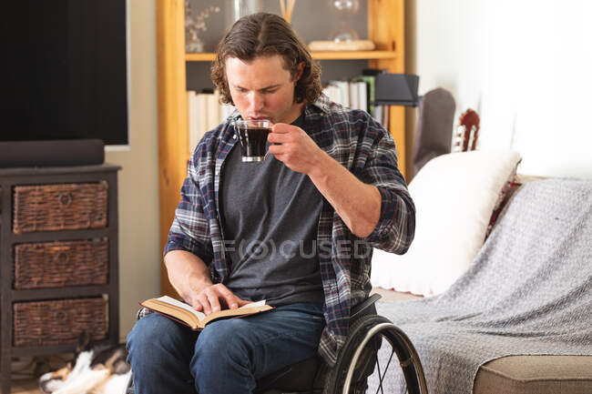 Hombre caucásico discapacitado sentado en silla de ruedas bebiendo café y leyendo un libro en casa. concepto de discapacidad y discapacidad - foto de stock