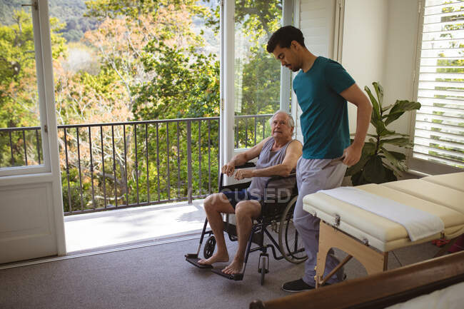 Fisioterapeuta masculino biracial tratando paciente masculino sênior em cadeira de rodas na clínica. cuidados de saúde seniores e tratamento de fisioterapia médica. — Fotografia de Stock