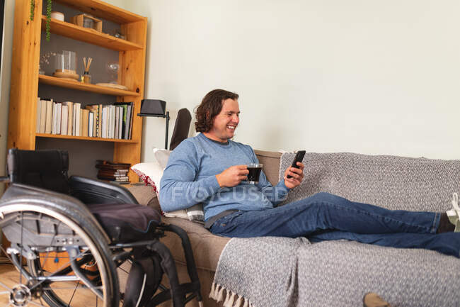 Homme handicapé caucasien tenant une tasse de café en utilisant un smartphone assis sur le canapé à la maison. handicap et handicap concept — Photo de stock