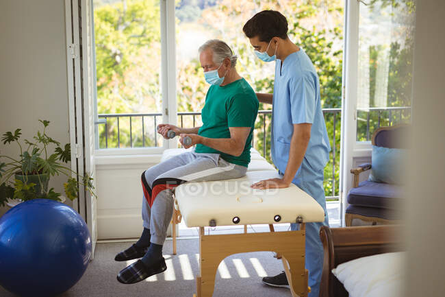 Biracial male physiotherapist with face mask treating arms of senior male patient at clinic. senior healthcare and medical physiotherapy treatment. — Stock Photo