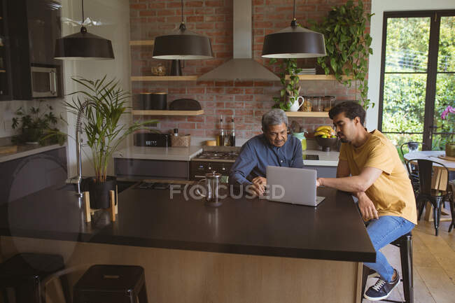 Biracial adult son and senior father using laptop in kitchen. family time at home together. — Stock Photo