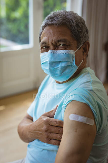 Portrait d'un homme âgé biracial portant un masque facial montrant un bandage sur le bras après une vaccination covide. soins de santé et mode de vie pendant la pandémie de covide 19. — Photo de stock