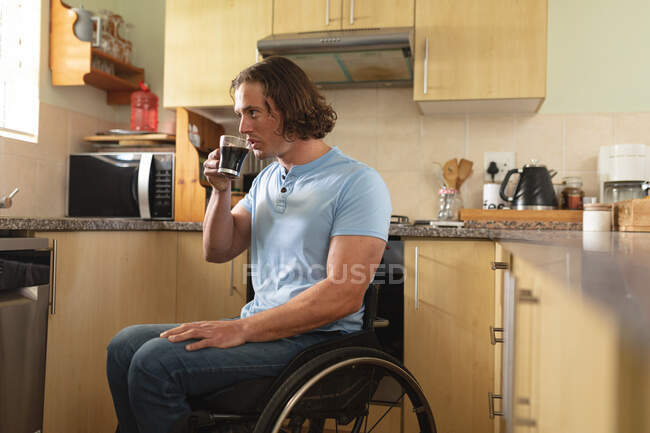 Кавказский инвалид, сидящий на инвалидной коляске и пьющий кофе дома на кухне. Концепция инвалидности и инвалидности — стоковое фото
