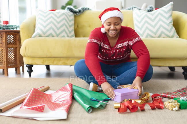 Feliz afro-americano plus size mulher em Papai Noel embrulhando presentes em casa. Natal, festa e conceito de tradição. — Fotografia de Stock