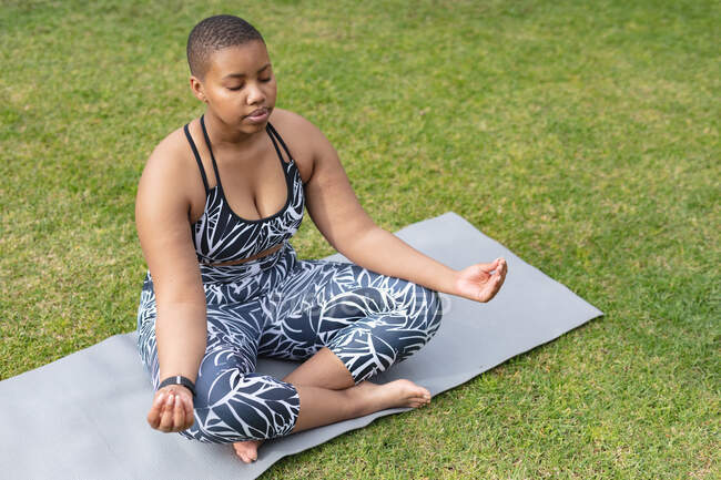 Focada afro-americana plus size mulher praticando ioga no tapete no jardim. fitness e estilo de vida saudável e ativo. — Fotografia de Stock