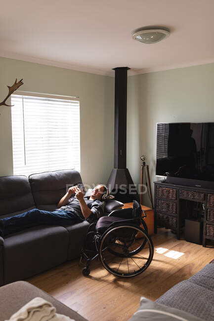 Hombre caucásico discapacitado con auriculares usando teléfono inteligente mientras está acostado en el sofá en casa. concepto de discapacidad y discapacidad - foto de stock