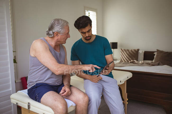 Біраціонний чоловічий фізіотерапевт використовує таблетку з пацієнтом старшого віку в клініці. лікувальна та медична фізіотерапія . — стокове фото