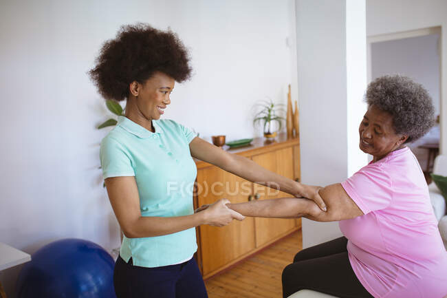 Lächelnde afrikanisch-amerikanische Physiotherapeutin, die Arme einer älteren Patientin in der Klinik behandelt. Senior Health und medizinische physiotherapeutische Behandlung. — Stockfoto