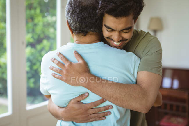 Sonriente hijo adulto birracial y padre mayor abrazándose en la sala de estar. tiempo en familia en casa juntos. - foto de stock
