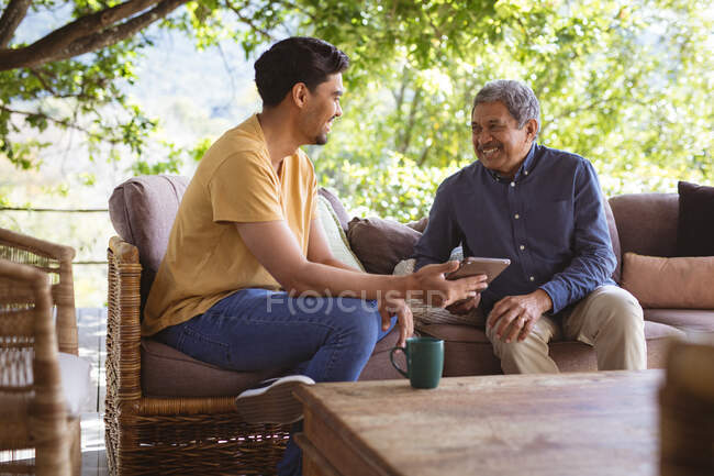 Sorrindo biracial filho adulto e pai sênior usando tablet no jardim. tempo de família em casa juntos. — Fotografia de Stock