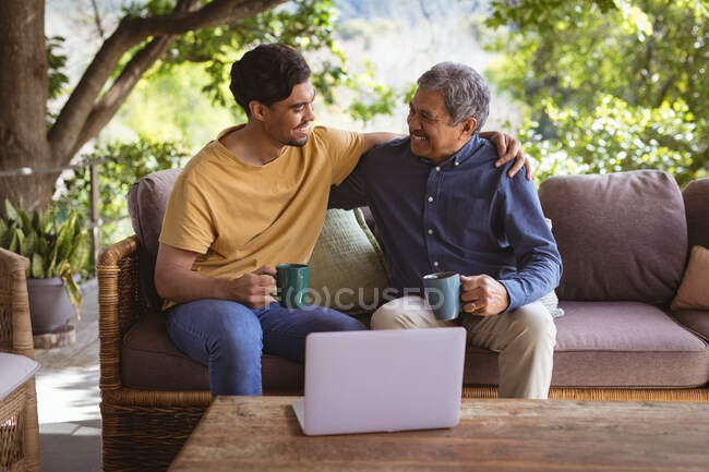 Sonriente hijo adulto birracial y padre mayor abrazando y bebiendo café en el jardín. tiempo en familia en casa juntos. - foto de stock