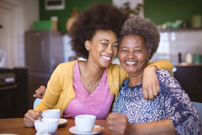 Portrait of smiling african american senior woman with adult daughter drinking coffee and embracing. family time at home together. — Stock Photo