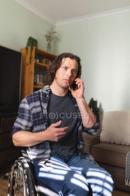 Caucásico hombre discapacitado sentado en silla de ruedas hablando en el teléfono inteligente en casa. concepto de discapacidad y discapacidad - foto de stock