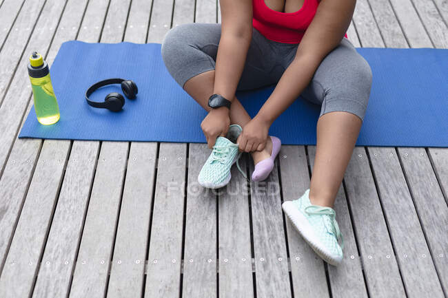 Sección media de mujer afroamericana de talla grande en ropa deportiva sentada en una esterilla de yoga y atando zapatos. fitness y estilo de vida saludable y activo. - foto de stock
