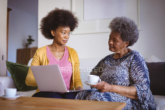 Mujer mayor afroamericana con hija adulta usando laptop. tiempo en familia en casa utilizando la tecnología juntos. - foto de stock