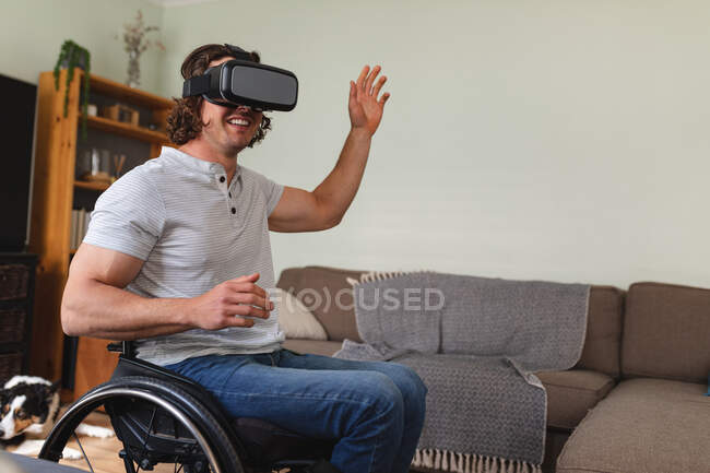 Uomo disabile caucasico seduto sulla sedia a rotelle indossando auricolare vr e gesticolando a casa. concetto di disabilità e handicap — Foto stock