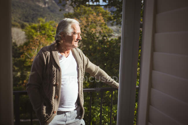 Hombre caucásico mayor mirando hacia el balcón en un día soleado. pasar tiempo en casa solo. - foto de stock