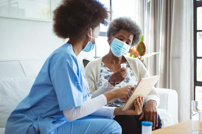 African american female doctor with face mask giving senior female patient pills at home. healthcare and lifestyle during covid 19 pandemic. — Stock Photo