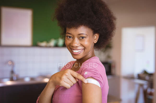 Retrato de una mujer afroamericana sonriente que muestra vendaje en el brazo después de la vacunación covid. salud y estilo de vida durante la pandemia de covid 19. - foto de stock