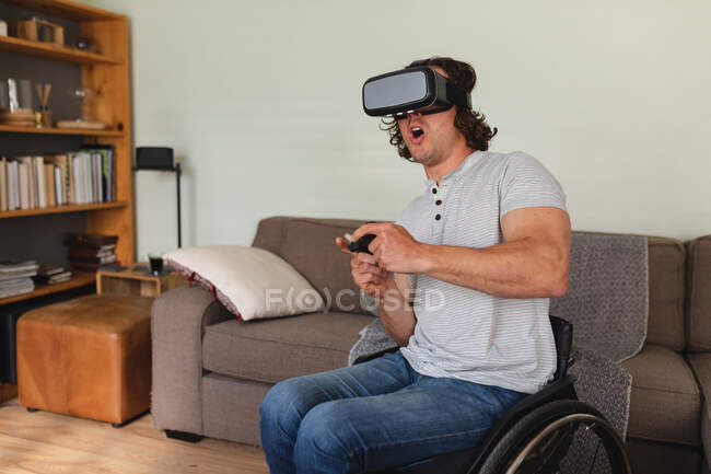 Caucasian disabled man sitting on wheelchair wearing vr headset and playing video games at home. disability and handicap concept — Stock Photo