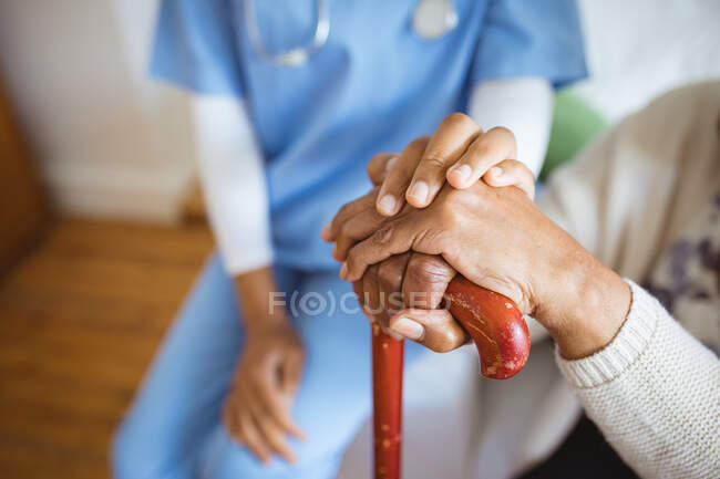Médica afro-americana de mãos dadas por uma paciente idosa em casa. cuidados de saúde e estilo de vida durante a pandemia de 19 pessoas. — Fotografia de Stock